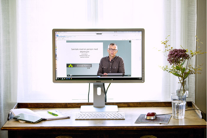 PC-skjerm som viser bilde av en mann som snakker. PC står på et skrivebord med blomst og vannglass,