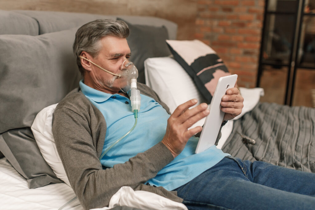 Mann som ligger op sofaen med en ipad i hånden og har på seg en forstøvermaske.