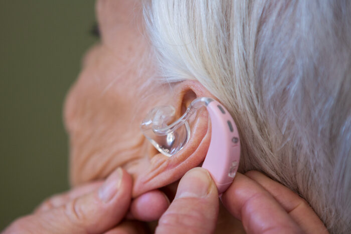 Bilde av en eldre dame og to hender som plasserer et høreapparat i øret hennes.