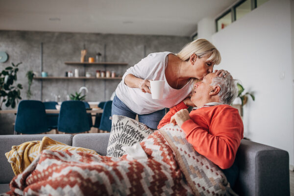 Fotografi av eldre par. Kvinnen kysser mannen, som ligger på sofaen med pledd, på pannen.