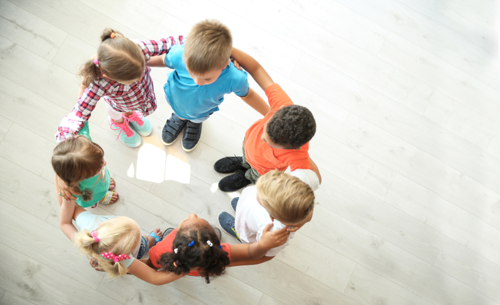 Little,Children,Making,Circle,With,Hands,Around,Each,Other,Indoors, Fotografi av barn som holder hverandre på skuldrene, i en sirkel. Fugleperspektiv/ovenfra