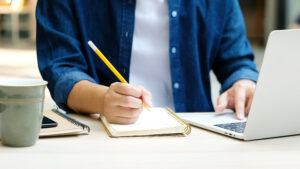 Bilde viser en person som sitter å skriver på en blokk med en pc ved siden av seg
