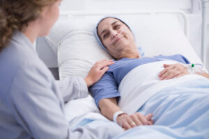 Syk og sengeliggende kvinne som smiler til helsearbeider som sitter ved sengen.
