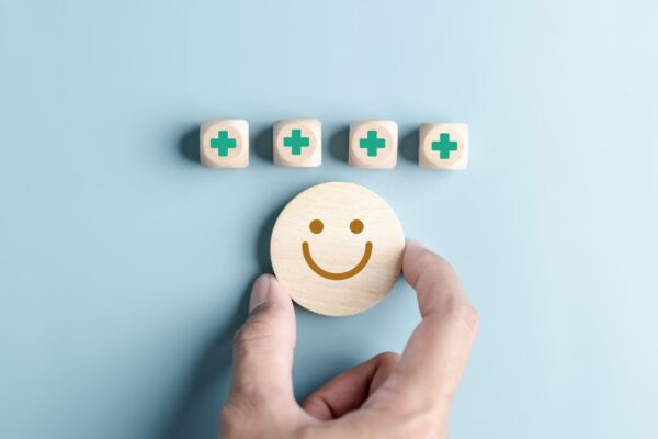Blå bakgrunn, en hånd som stiller frem et smilefjes i tre, plassert under terninger med "helsekors" symbolet på.