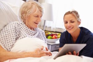 Ung kvinnelig helsepersonell, snakker med eldre dame som sitter i seng med ipad