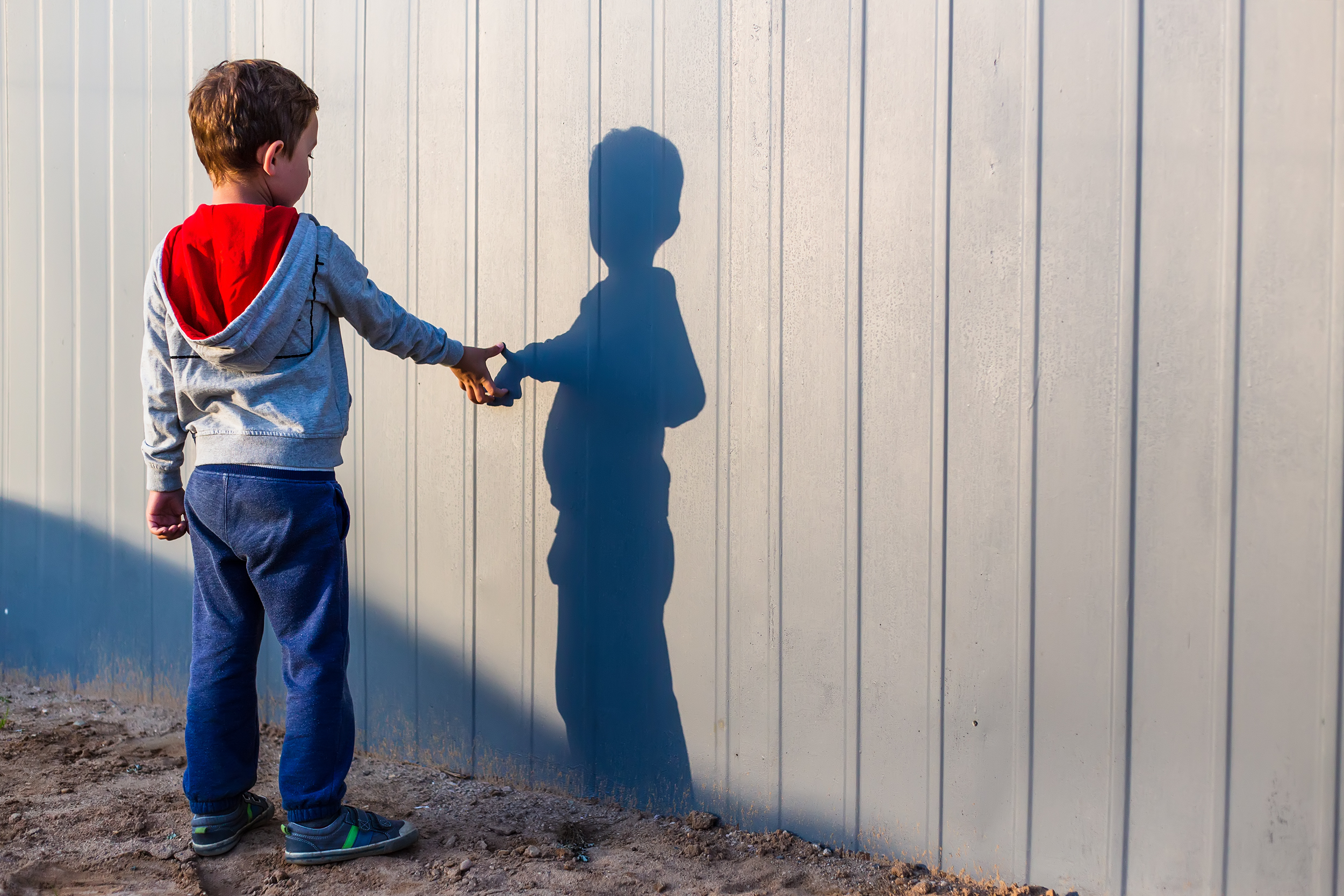 Boy,And,His,Shadow.,Lonely,Little,Child,Playing,With,His Fotografi av barn stående foran et gjerde, nysgjerrig på sin egen skygge