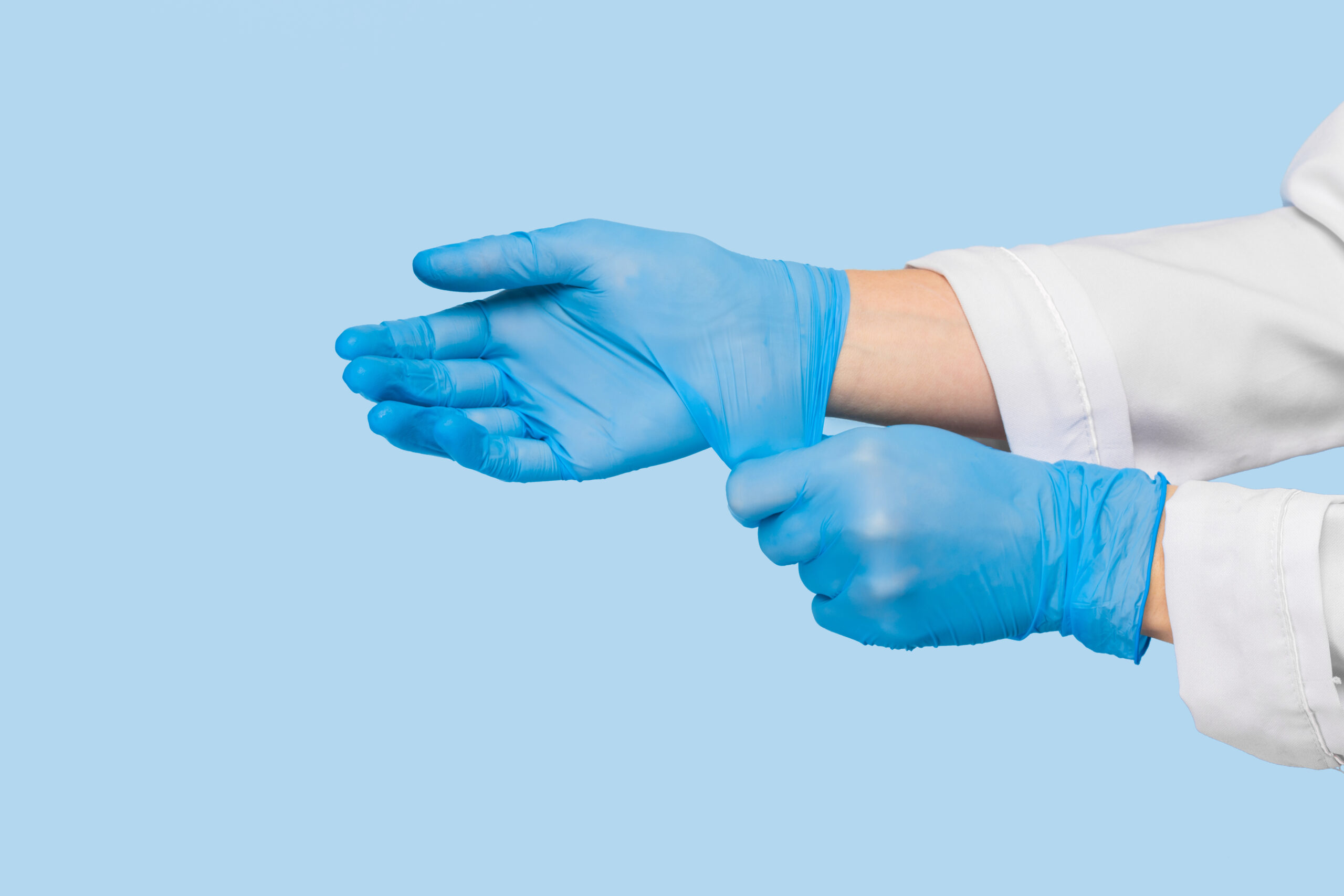 The,Doctor,Puts,On,Sterile,Gloves,On,A,Blue,Background. Foto av lege som tar på seg hansker, med fokus på smittevern. Blå bakgrunn, blå hansker, hvit legefrakk