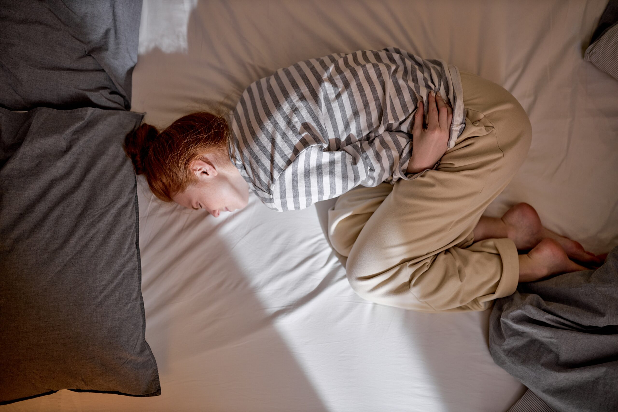 Top,View,Of,Sick,Young,Ginger,Redhead,Woman,Lying,In Fotografi av ung kvinne som holder seg på magen, liggende i forsterstilling i senga