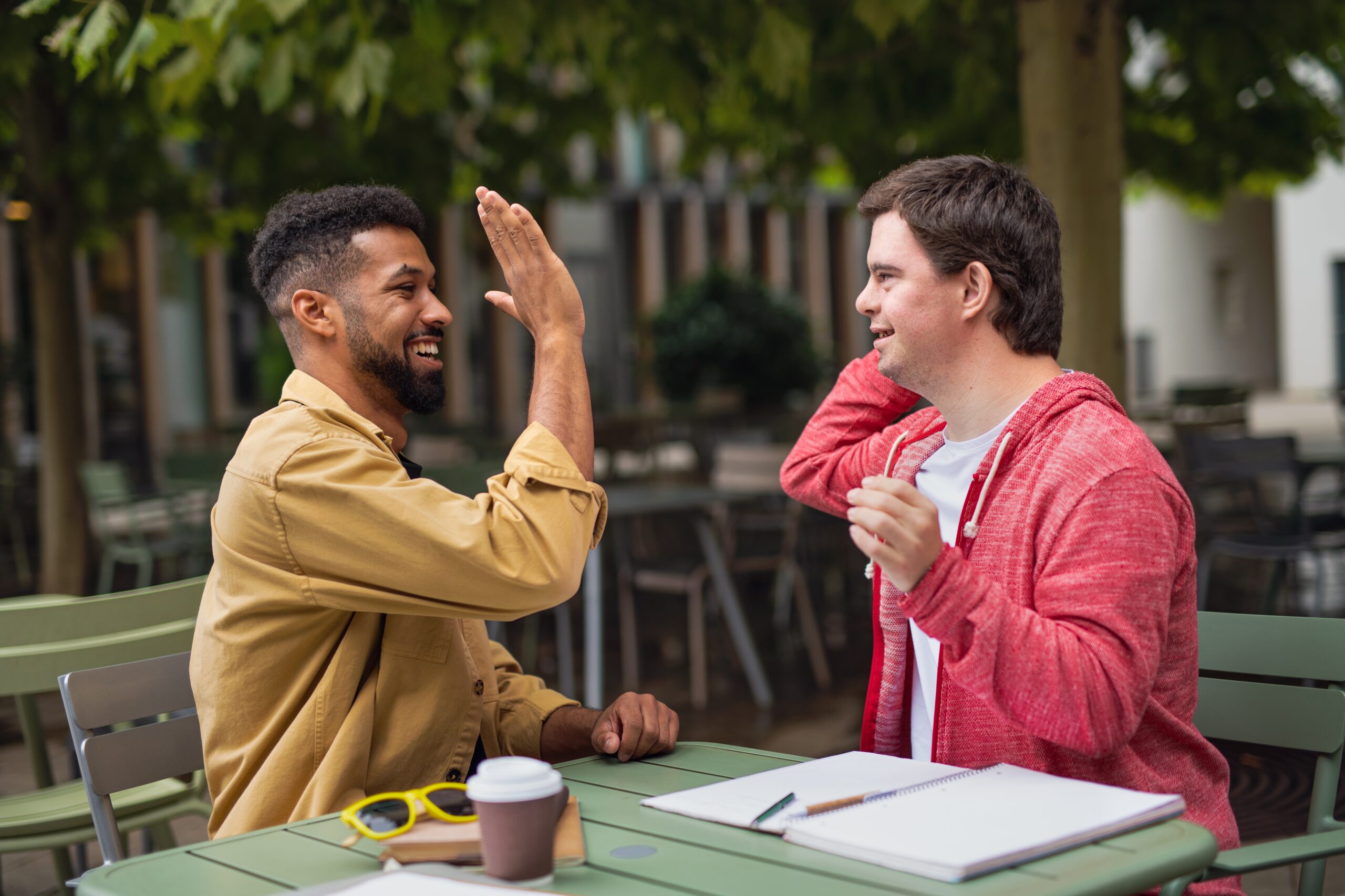 Young,Man,With,Down,Syndrome,With,Mentoring,Friend,Sitting,Outdoors Fotografi av to menn som high-five'er hverandre. En av dem har downs og har på seg en rød genser. Den andre personen har på seg en gul skjorte. De sitter ute.