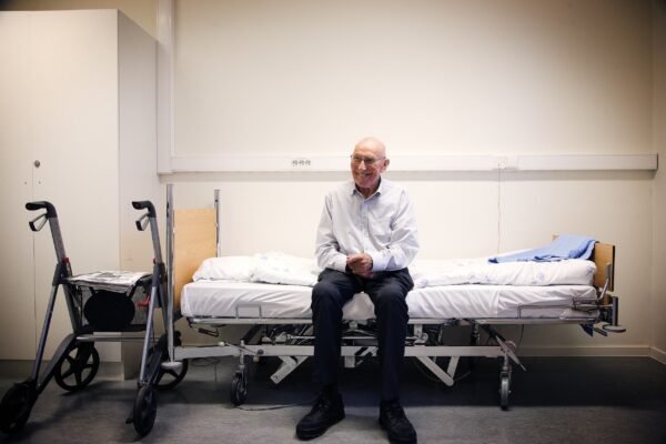 Fotografi av eldre mann, sittende på kanten av en sykehusseng. Blid og smilende