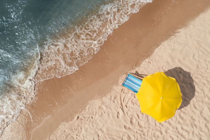 Fotografi tatt i fugleperspektiv av gul parasoll og blå solstol på en strand.