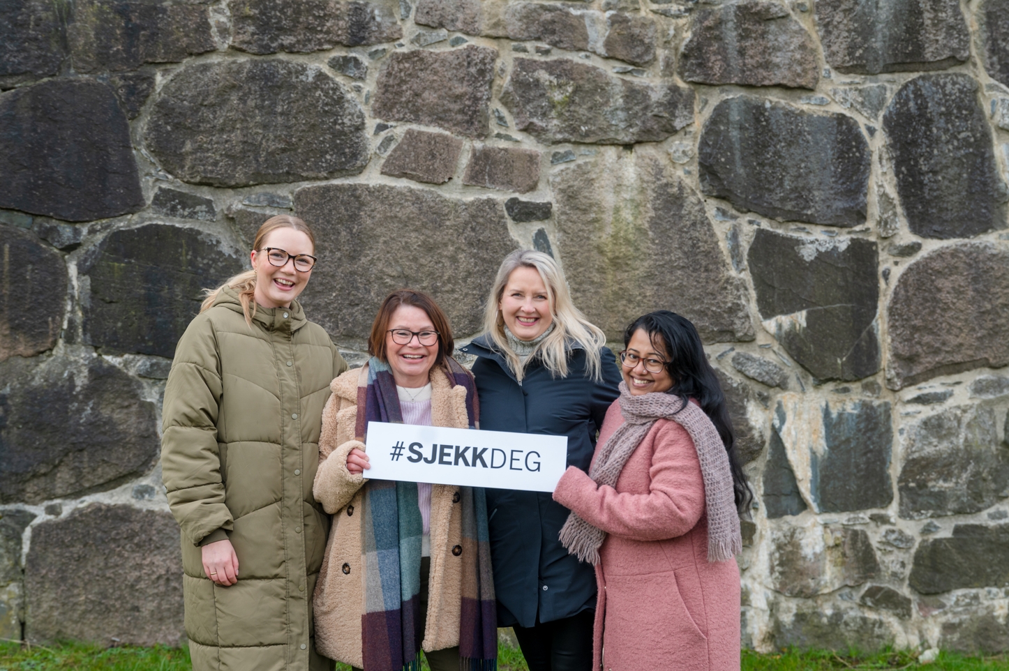 Sjekk-deg- fotografi av tre kvinner stående foran en murvegg med skiltet: sjekk deg
