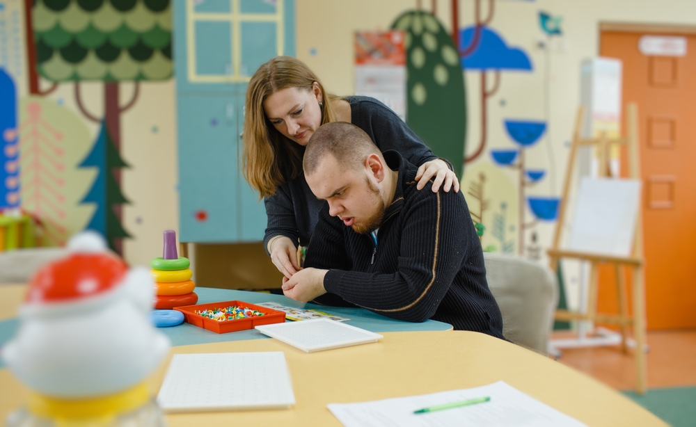 A,Female,Psychologist,Works,With,An,Adult,Boy,With,Autism Fotografi av person med utviklingshemming i situasjon med lærer / helsepersonell, sittende ved en pult