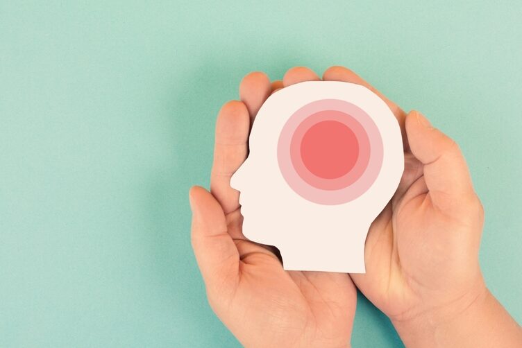 Human,Brain,,Migraine,Headache,Attack,,Psychological,Pressure,And,Stress,,Mind Bildeillustrasjon med fokus på traume. To hender holder på et menneskehode-utklipp i profil, med et rødt verkende merke i hjernen