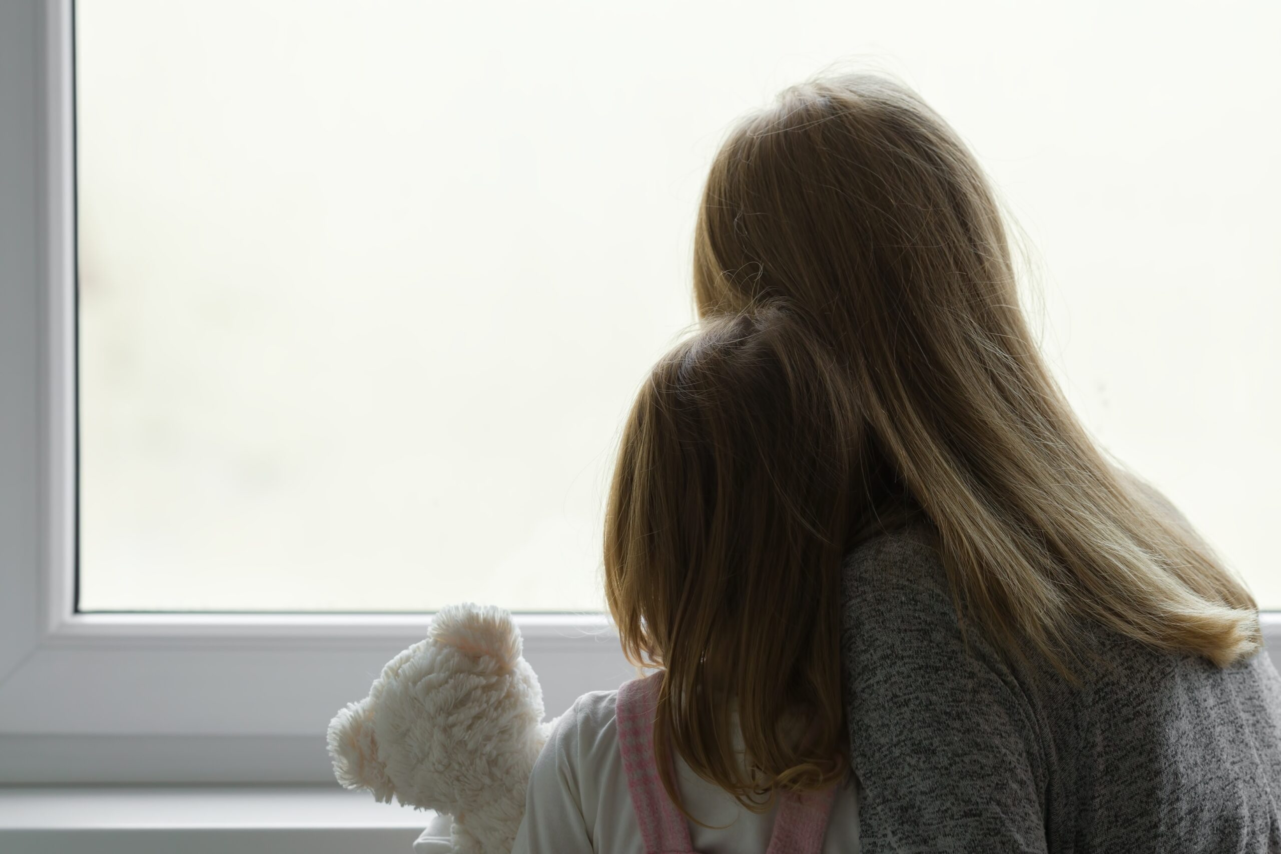 Little,Girl,Holding,White,Teddy,Bear.,Young,Adult,Mother,And Fotografi av silhuetten / ryggene / skyggene til mor og datter som titter ut av et vindu. Jenta holder en bamse