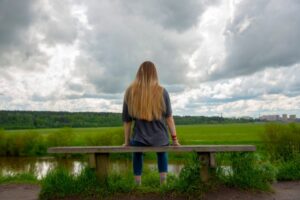 Bilde av ung kvinne sittende med ryggen vendt mot kamera på en benk. I bakgrunnen er det en grønn ås og mørke skyer på himmelen hvor solen bryter litt igjennom.
