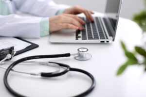 Illustrasjonsbilde av helsepersonell i hvit som sitter ved bord og skriver på en laptop. På bordet ligger også et synlig stetoskop.