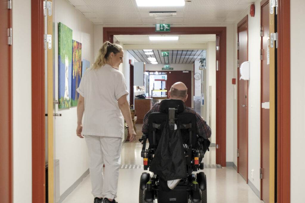 Vi ser bakfra helsearbeider og mann i rullestol sammen på veg nedover en korridor.