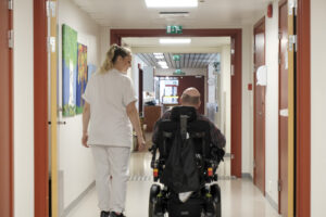 Vi ser bakfra helsearbeider og mann i rullestol sammen på veg nedover en korridor.