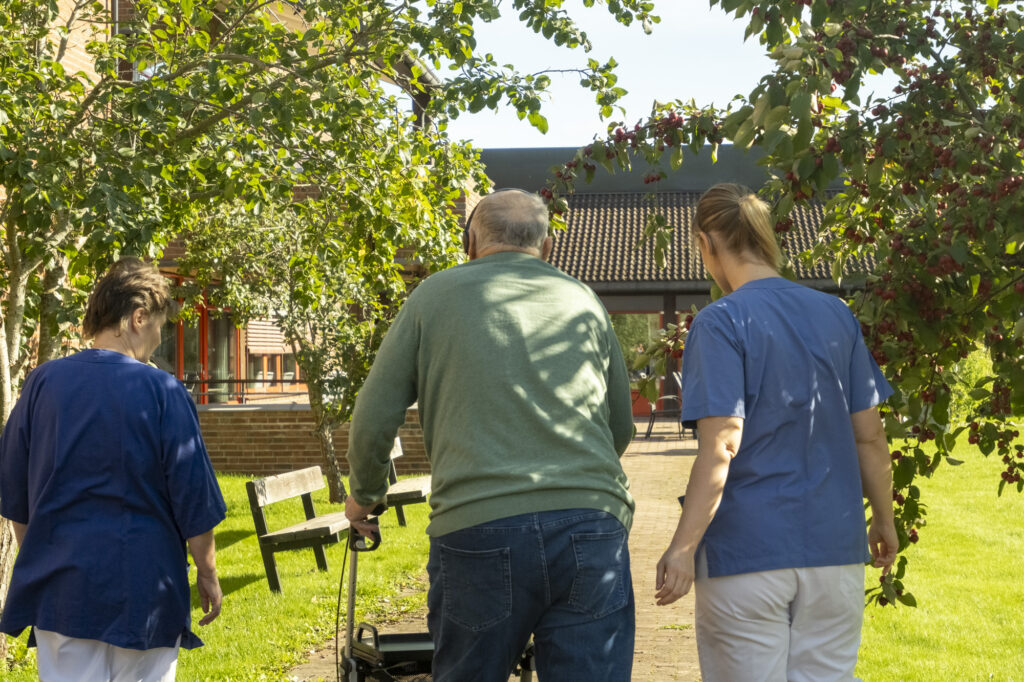 Eldre mann med rullator spaserer sammen med en helsearbeider på hver side av seg.