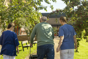 Eldre mann med rullator spaserer sammen med en helsearbeider på hver side av seg.