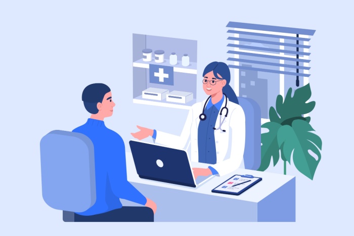 Kvinnelig lege sitter bak et skrivebord inne på et kontor med PC foran seg og snakker med pasient som sitter å stol på andre siden av skrivebordet