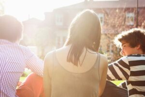 Illustrasjonsbilde av en voksen kvinne sittende mellom to tenåringsgutter med ryggen vendt mot kamera. I Bakgrunnen skimter man solskinn og en boligblokk