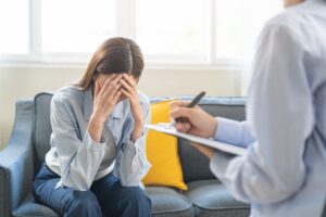 Bilde viser en dame som sitter med hode lent framover med hendene over ansiktet. I tillegg ses litt av en person som sitter å noterer på en blokk.