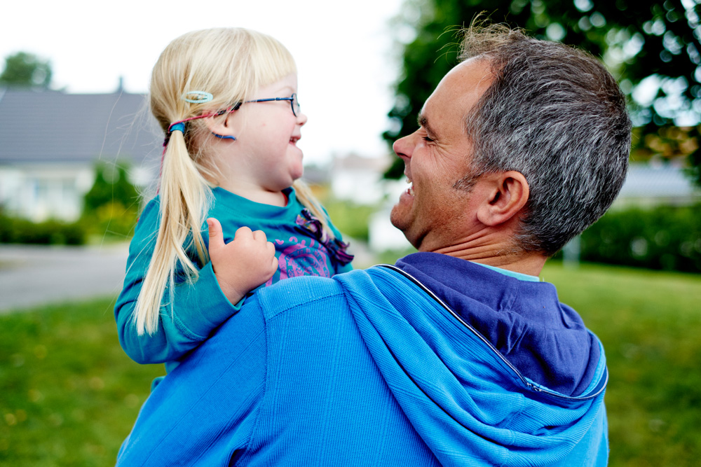 Illustrasjonsbilde, Sykehuset i Vestfold. FOTO: MORTEN RAKKE fotografi av mann som bærer barn, utendørs på plenen. Mannen har blå jakke, og jenta har lyst hår og briller
