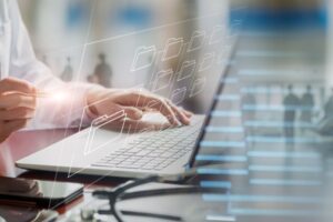 Bilde av deler av hvitkledd person sittende forran pc. På skrivebordet ligger et stetoskop. Ment å illustrere lege som henviser elektronisk.