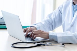 Illustrasjonsbilde av lege sittende forran pc med stetoskop liggende ved siden av.