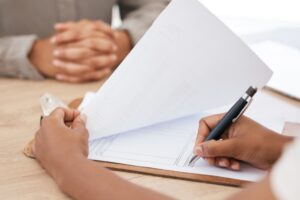 Foto med fokus på helsepersonell som skriver på et skjema. I bakgrunnen sitter en person med foldede hender, ment å illustrere en pasient.