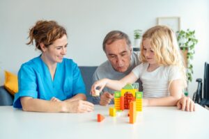 Kvinnelig helsepersonell sammen med eldre pasient og hans barnebarn som leker med byggeklosser.