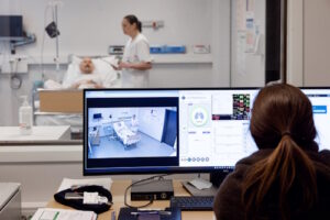 Person som overvåker skjerm og inn på pasientrom hvor det står en pleier som steller en i sengen.