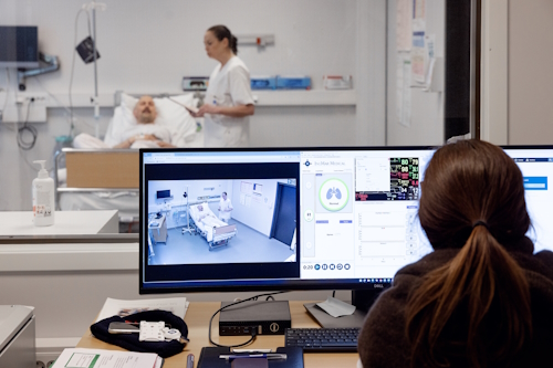 Person som overvåker skjerm og inn på pasientrom hvor det står en pleier som steller en i sengen.