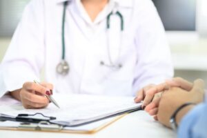 Illustrasjonsbilde av lege i samtale med pasient hvor bilde har fokus på pasientens hender og legens overkropp. Legen skriver på et papir, ment å illustrere en legekonsultasjon.