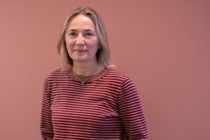 Fotografi av Anna-Brita knapskog i forbindelse med podkast-innspilling. Stående foran beige vegg