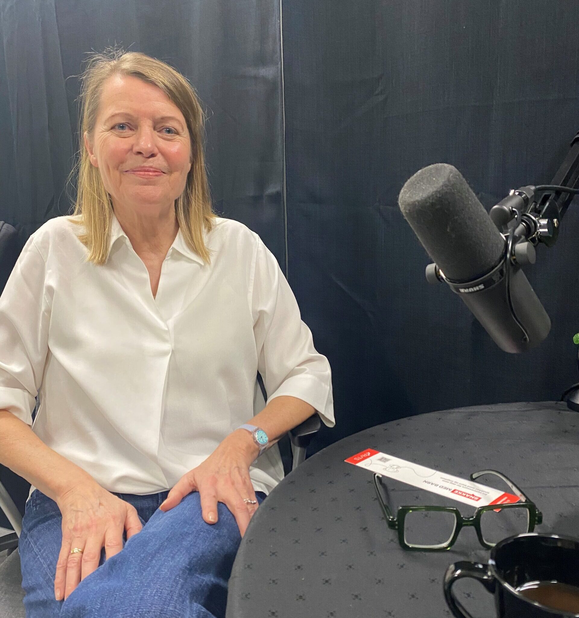 Podkastgjest Ane H. Simonsen sittende i studio, foran en sort vegg med mikrofonen pekende mot seg. Fotografi