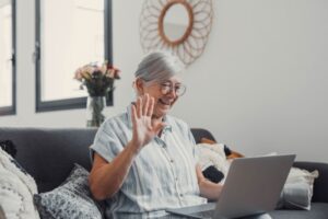 Eldre damen som sitter i sofaen og smiler og vinker til en dataskjerm som hun har på fanget