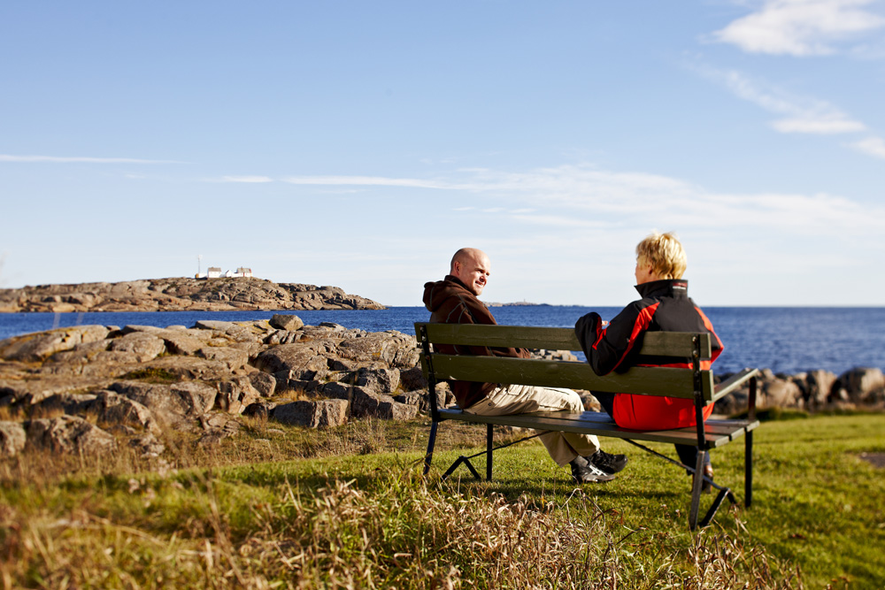 Fotografi av to mennesker sittende på en benk ved havet i solen