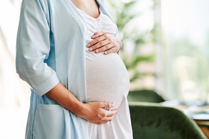 Bilde av gravid kvinne som holder seg rundt magen fra skuldrene og ned. I bakgrunnen ses noen planter.