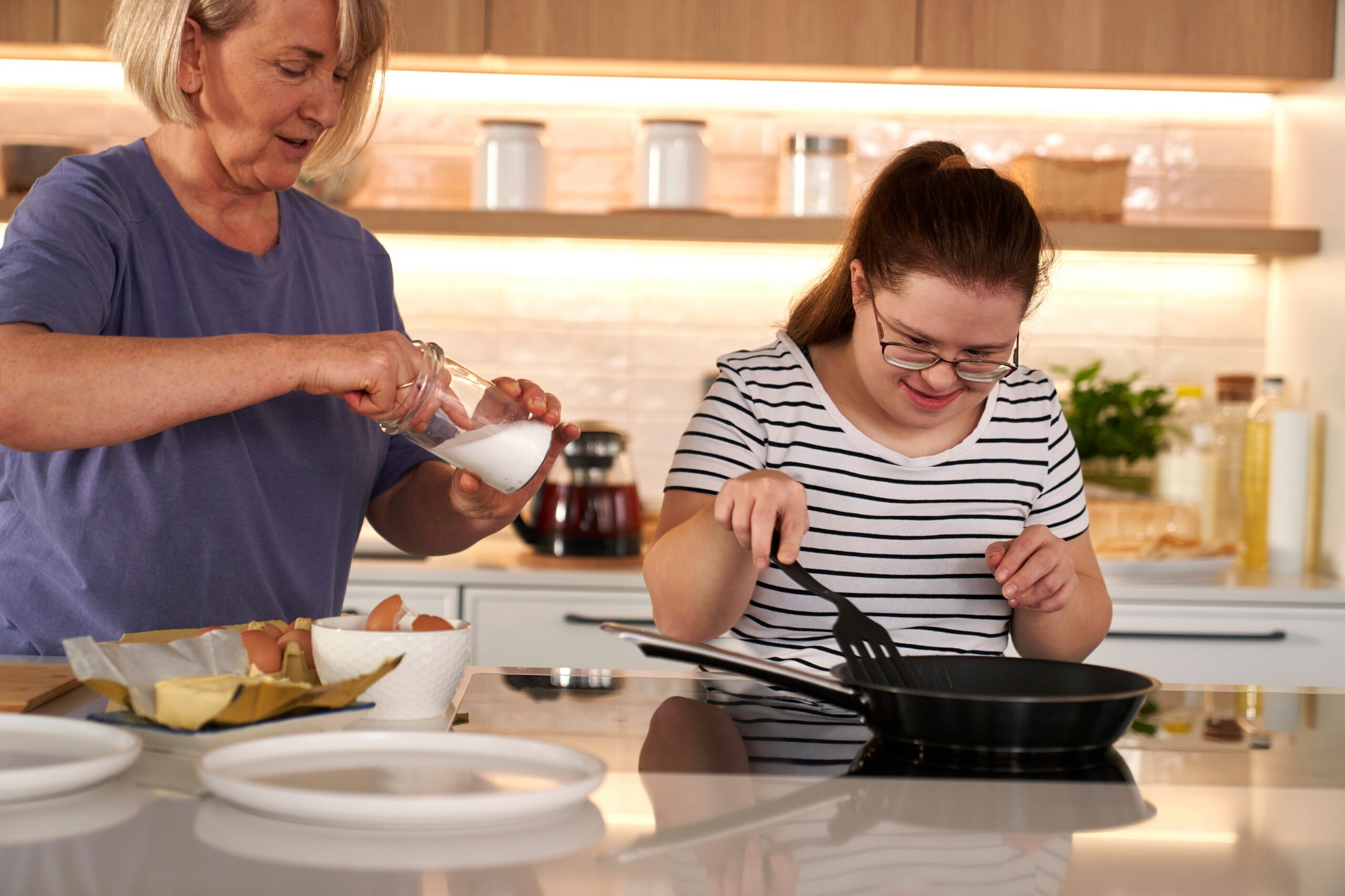 Ung person med downs-syndrom forbereder et måltid med en voksen person stående ved siden av. Foto