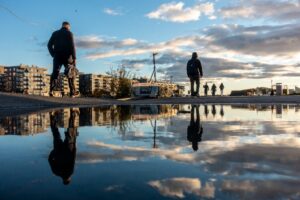 Flere personer går langs en brygge i Bjørvika ved solnedgang, med bygninger i bakgrunnen og speilinger i en vannpytt i forgrunnen