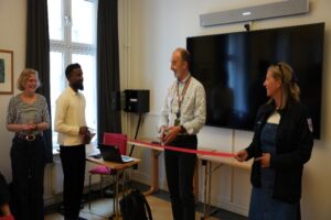 Fire personer står i et møterom under en markering med rødt bånd. En person i midten holder en saks, mens de andre står rundt. I bakgrunnen er det et stort TV-skjerm, vinduer og et lite bord med en bærbar PC. Foto.