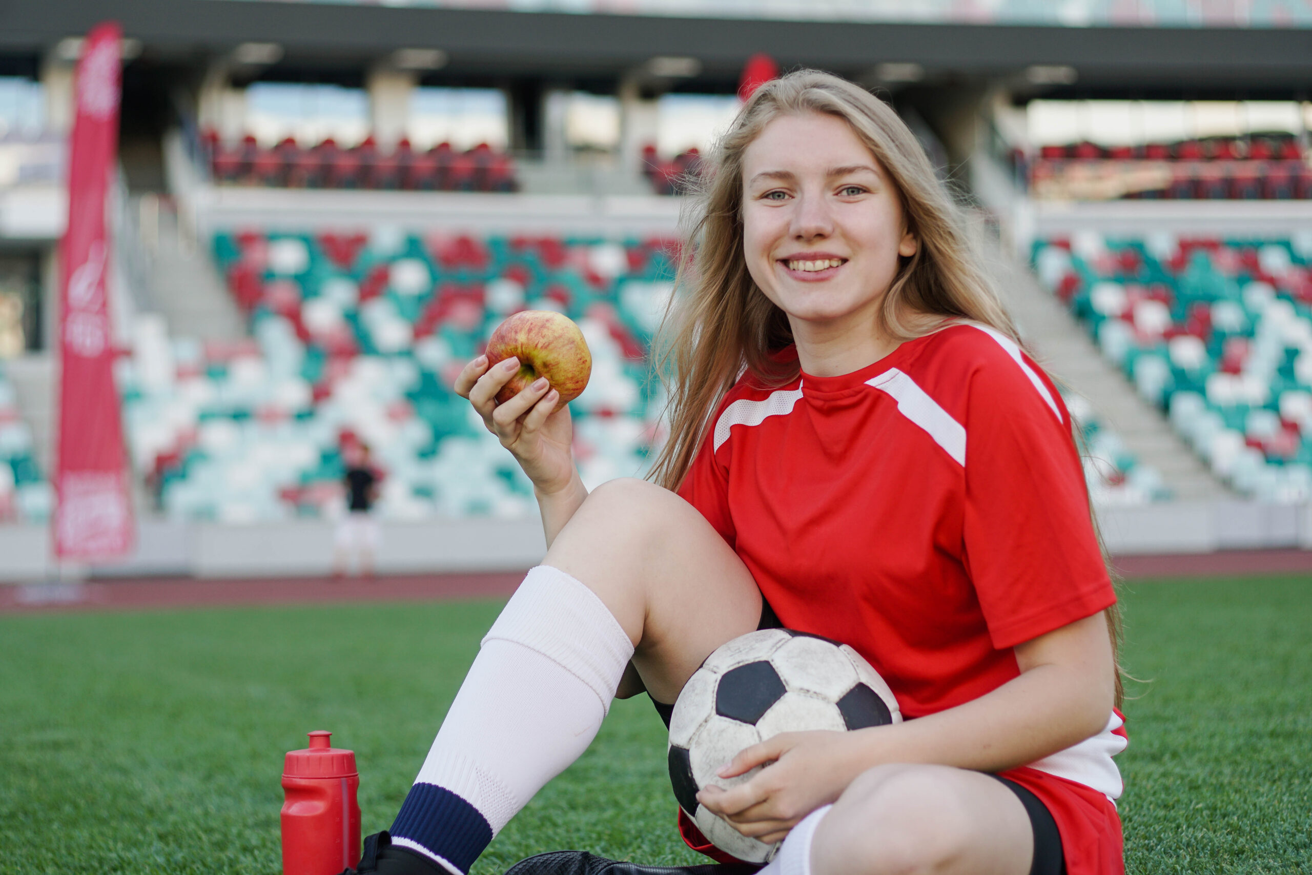 Portrett av en ung kvinnelig fotballspiller som sitter på fotballbanen med et eple etter trening. Foto