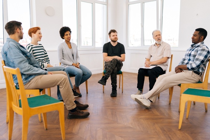 Wide shot of diverse people sitting in circle, sharing personal problems at psychotherapy session. Mature male psychologist therapist helping multiracial younger and older patients at group therapy.