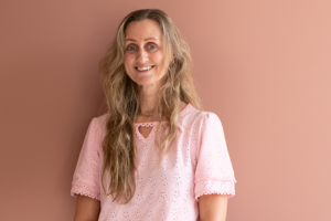 Podkast-gjest Lene Guttulsrud stående foran en beige vegg, smilende. Foto